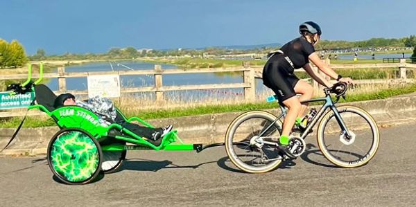 Cyclist pulling a green cart with a child by a lakeside road.