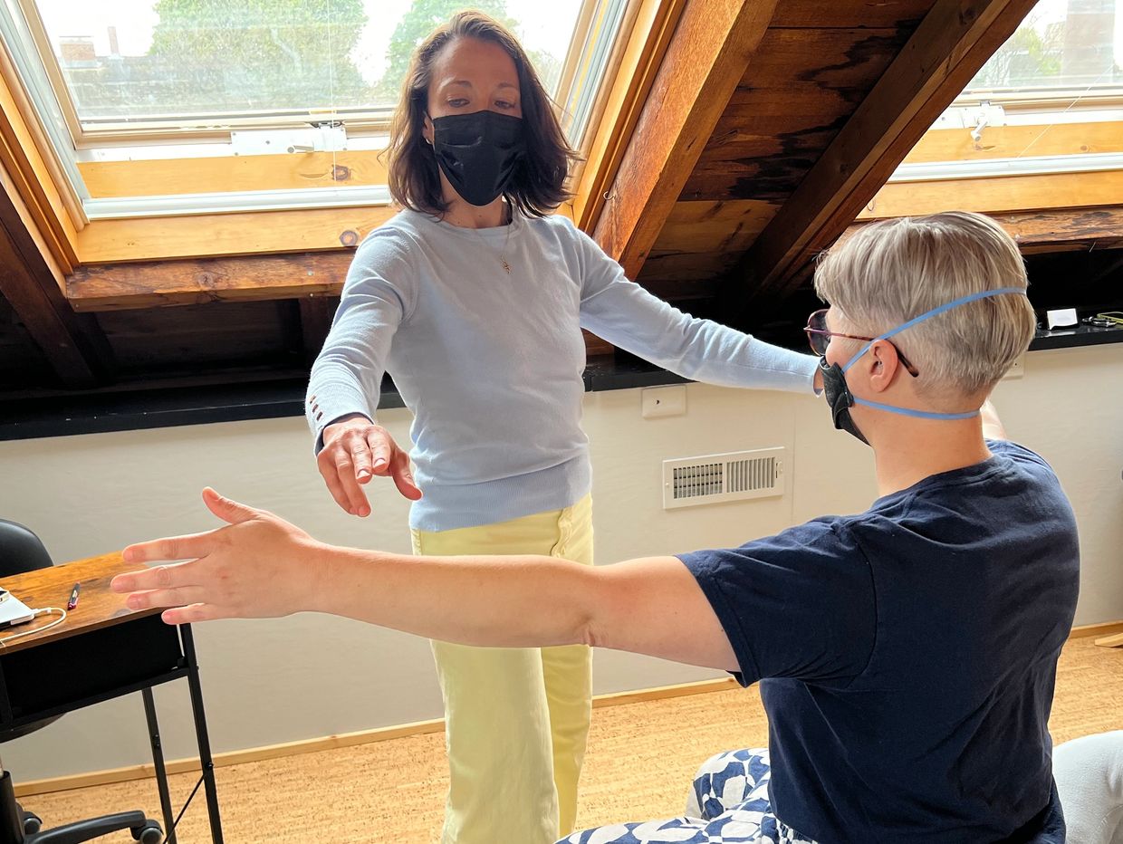 Two masked individuals in a bright room, engaged in a stretching or exercise activity.