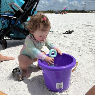 A toddler playing with a purple bucket and blue shovel on the beach.