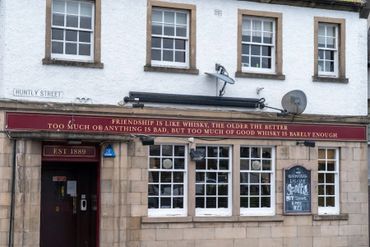 Pub facade with a witty whisky-themed friendship quote and a street sign of Huntly Street.