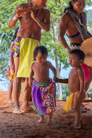 Indigenous adults play flute and drum while children dance in traditional attire.