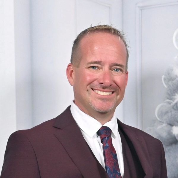 Smiling man in a suit and patterned tie against a light background.