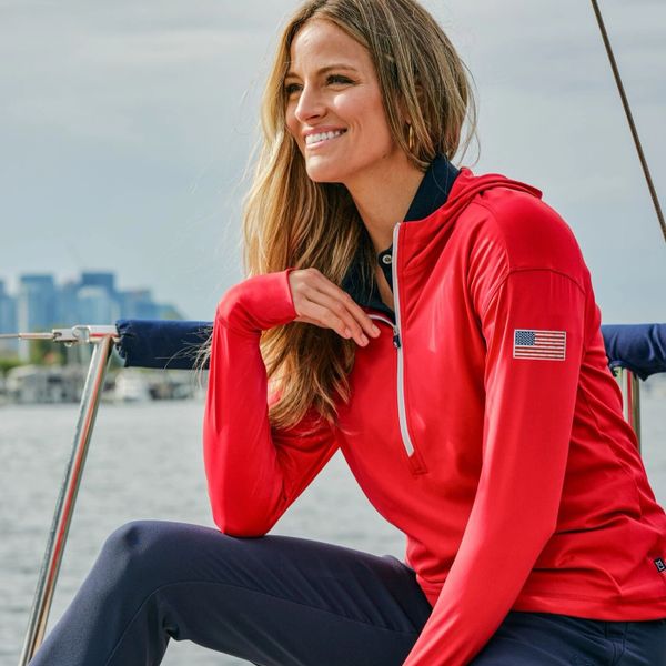 Smiling woman in a red jacket with an American flag patch, sitting on a boat by the water.