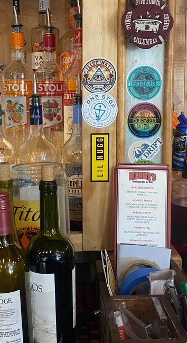 Bar shelf with various liquor bottles and brewery stickers on a wooden panel.