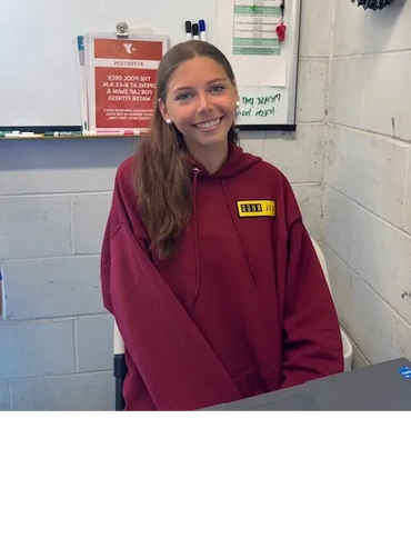 Smiling young woman in maroon hoodie sitting at a desk in a casual room.