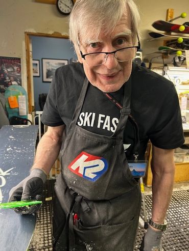 Elderly man in a workshop holding a ski, wearing a K2 apron and gloves.