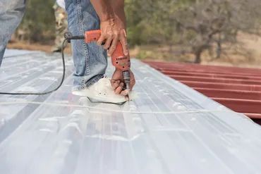 Person using a power drill on a metal roof panel outdoors.