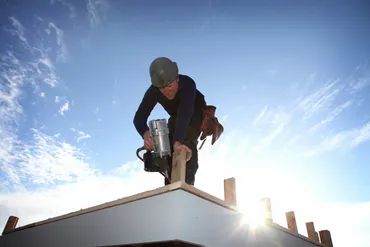 Worker using nail gun on rooftop under a bright sky.