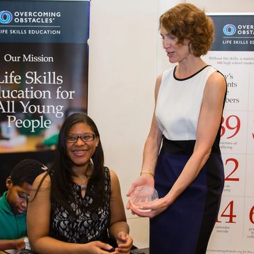 Johileny smiles as she receives a glass award from Jill a tall white woman with short hair.