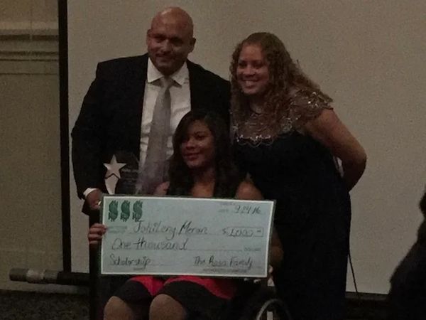 Johileny holds a big check. and smiles to the camera behind her a latinx woman and man.