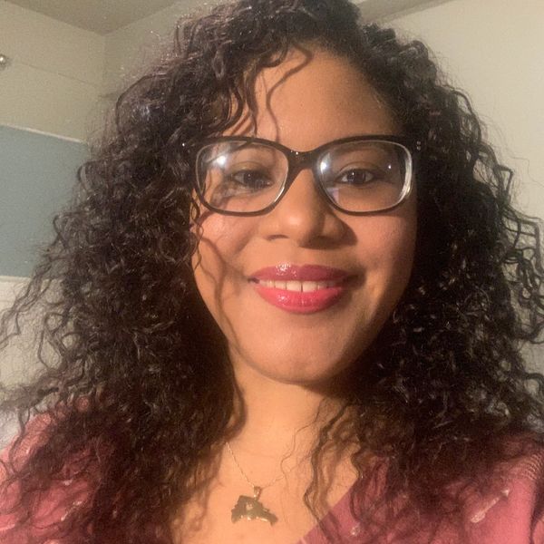 Portrait of Johileny with big curly hair. She smiles, wears glasses, and a necklace DR icon.