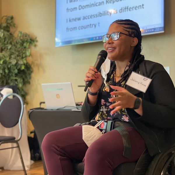 Johileny, speaks into a microphone at a conference. She wears glasses & has cornrows.