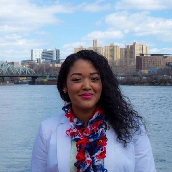Portrait of Mariela with NYC buildings behind. Curly hair to one side, blouse, and red/blue scarf.