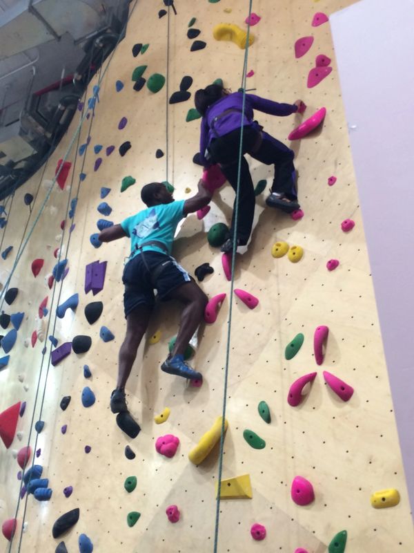 Johileny climbs up a indoor rock climbing wall. Beside her a black man supporting the clibming.