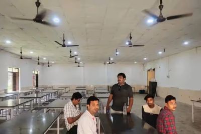 Five men sitting and standing in a large, empty cafeteria room with ceiling fans.