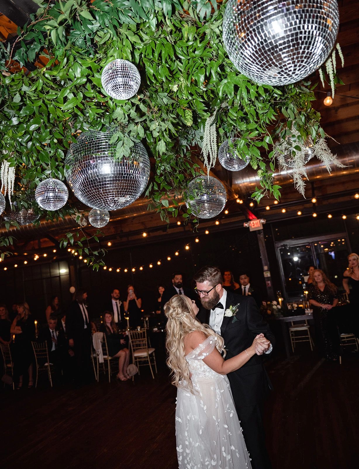 Bride and groom dance under a disco ball and fresh greenery installation. 