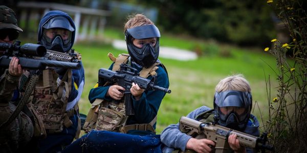 Children wearing tactical gear and masks playing paintball outdoors.
