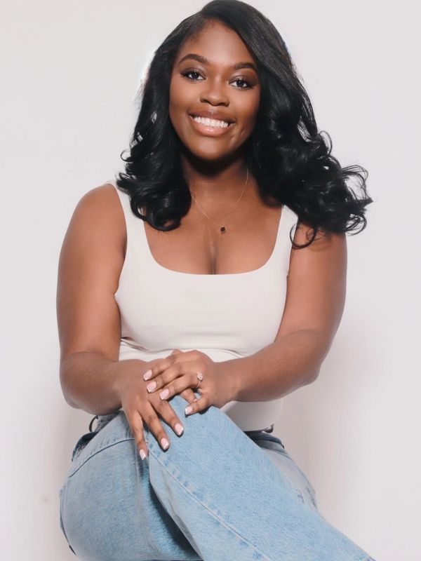 Smiling woman with wavy black hair, wearing a white tank top and blue jeans, sitting relaxed.