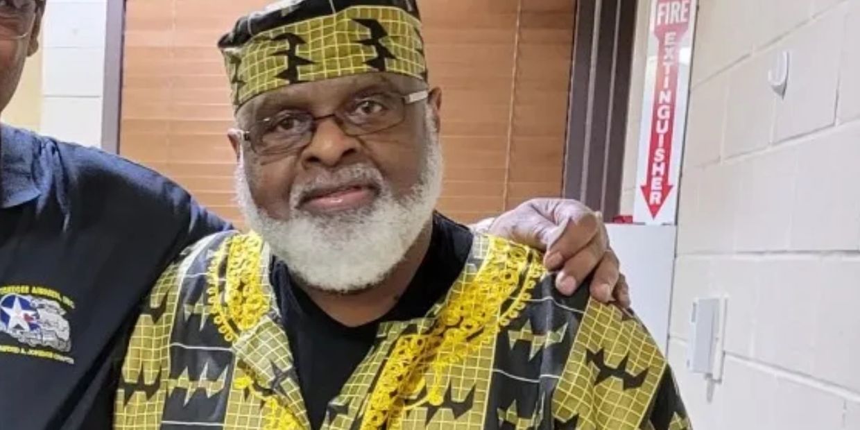 Elderly man in traditional African attire with a white beard and glasses.
