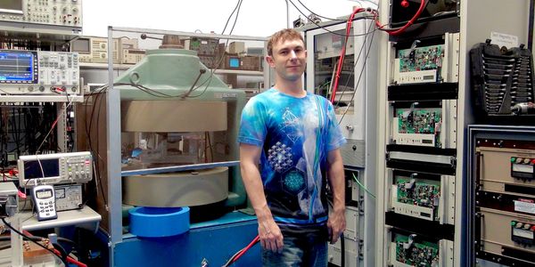 A man in a blue patterned shirt stands in a high-tech laboratory surrounded by electronic equipment.