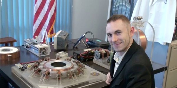A man in a suit smiles beside a scientific apparatus on a desk with an American flag in the background.