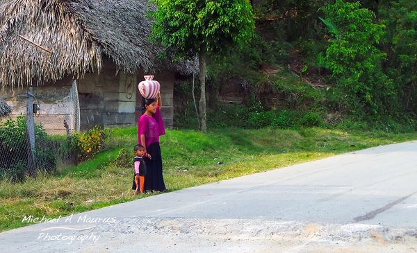 Maya woman standing on the side of the road with a vase on her head and holding a young child with h