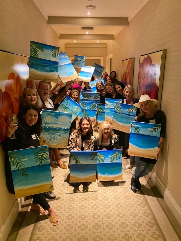 Group of women proudly displaying their beach-themed paintings in a hallway.