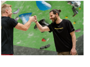 Two men in black shirts fist bump at an indoor climbing gym.