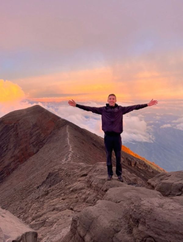 Young male tourist standing on top of Mount Agung, Bali at sunrise with arms spread wide.