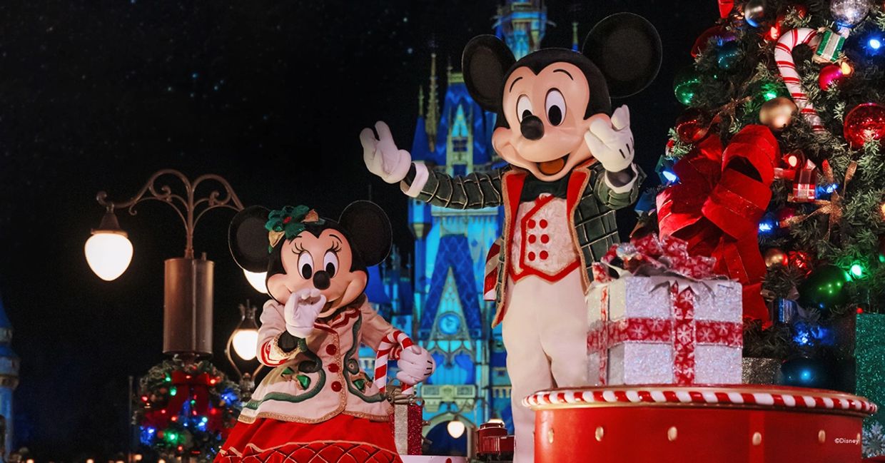 Mickey y Minnie Mouse vestidos con trajes navideños frente a un castillo iluminado.