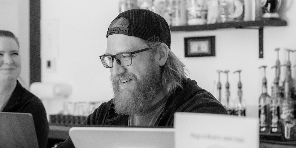 Bearded man with glasses and cap smiling while working on a laptop.