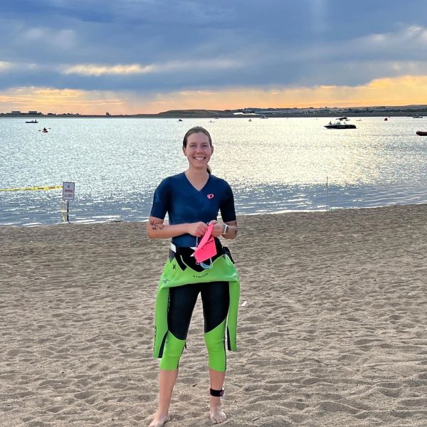 Woman smiling on the beach in wetsuit with water and boats behind.