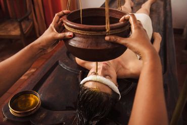 A woman receives an Ayurvedic oil treatment with oil dripping on her forehead.