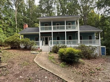 Two-story blue house with white railings surrounded by trees.
