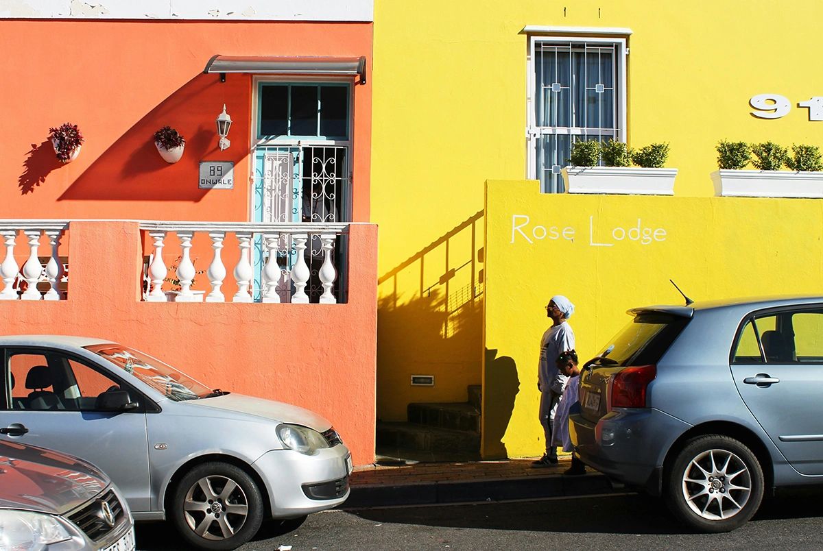 Vibrant street scene in Bo-Kaap, Cape Town, featuring brightly painted buildings in orange and yellow with white trim and decorative railings. A woman and child walk past “Rose Lodge” written on the yellow wall, while two parked cars line the street in front. The colorful facades and playful shadows highlight the neighborhood’s iconic charm. Vibrant orange and yellow Bo-Kaap houses with resident and parked cars, historic Malay Quarter featured in Cape Town guide
