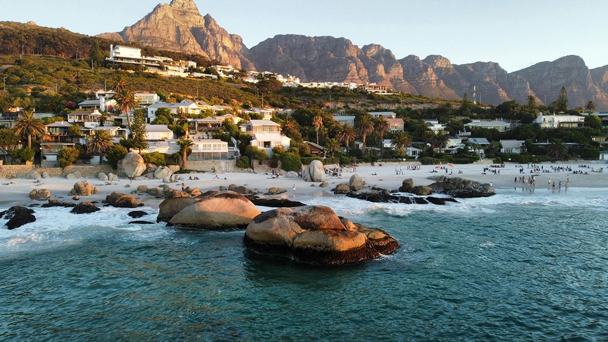 Aerial view of Clifton Beach in Cape Town at golden hour, with waves crashing against large boulders in the foreground and people relaxing along the white sandy shore. Upscale homes are nestled into the hillside, backed by the dramatic peaks of the Twelve Apostles mountain range glowing in the warm evening light. Clifton's granite boulders and luxury homes with Twelve Apostles mountain backdrop, exclusive beaches in Cape Town guide