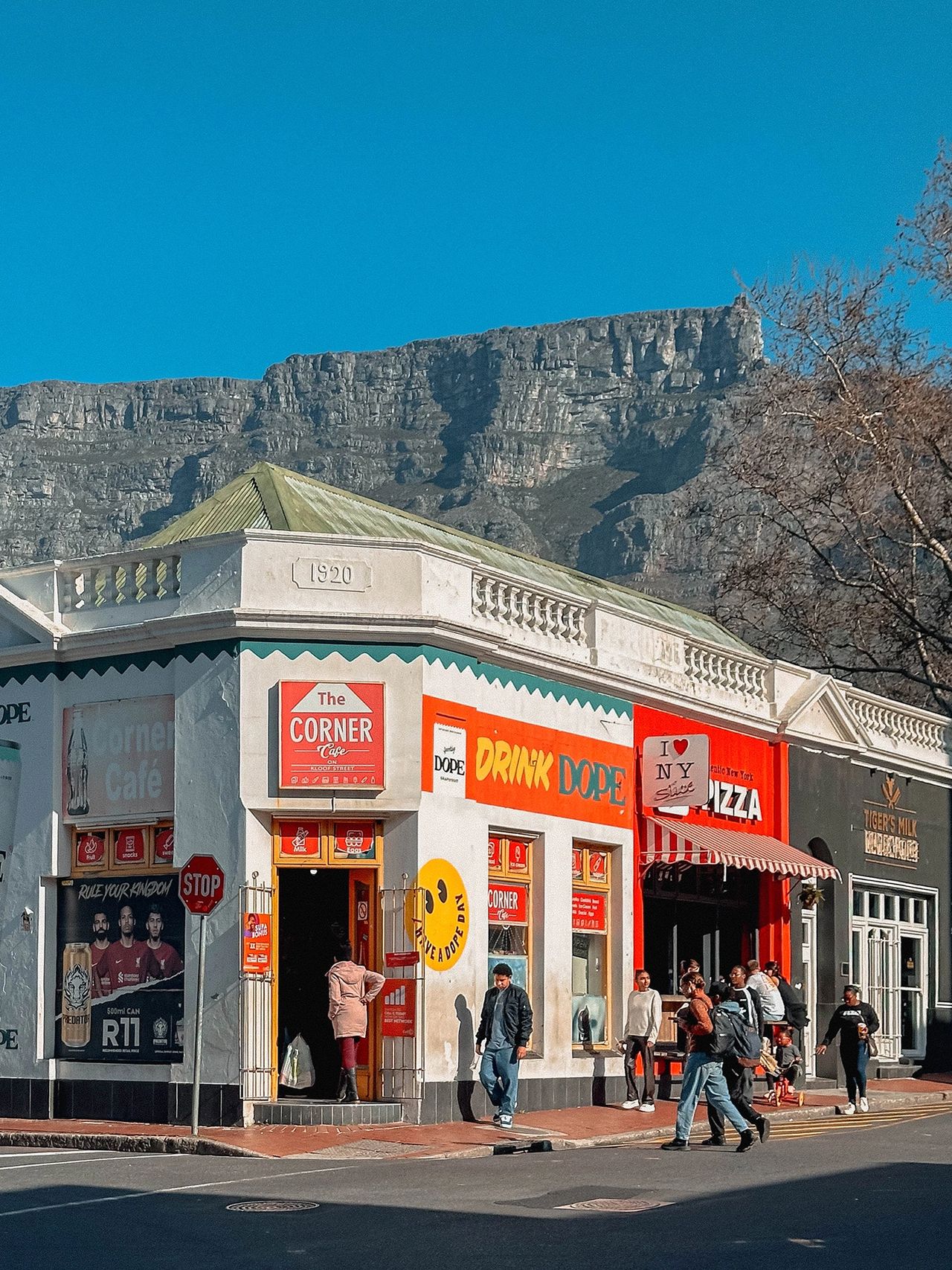 Kloof Street: Offers some of the best restraurants and coffee shops in the city Kloof Street's colorful Corner Café with Table Mountain backdrop. Text reads: "The Corner Café," "DRINK DOPE," "PIZZA," "TIGER'S MILK"