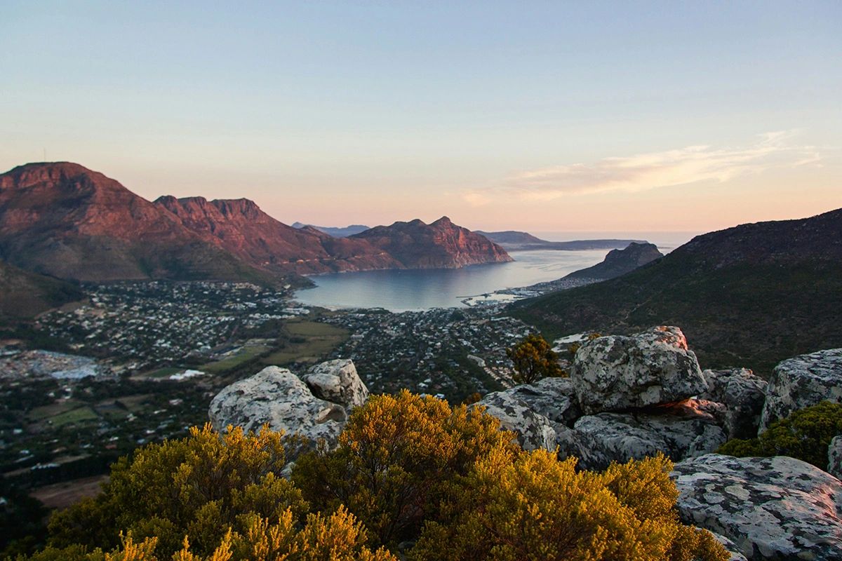 This stunning photo captures a panoramic view of Hout Bay at golden hour, one of Cape Town’s most picturesque and peaceful coastal enclaves. Surrounded by dramatic mountains and a tranquil bay, Hout Bay offers a slower pace, breathtaking hikes, and easy access to Chapman’s Peak Drive—one of the most scenic coastal roads in the world. It’s the perfect escape for nature lovers, photographers, and travelers looking to experience Cape Town beyond the usual tourist trail. Panoramic view of Hout Bay at sunset with mountains meeting the Atlantic Ocean, peaceful retreat in Cape Town guide