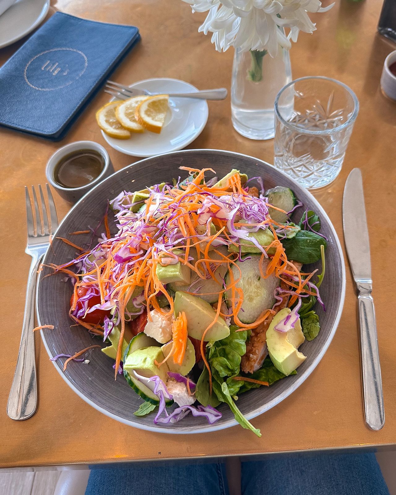 Colorful salad served in a gray bowl on a golden table, topped with shredded carrots, purple cabbage, avocado slices, cherry tomatoes, and grilled chicken. A small dish of dressing, a glass of water, a vase with white flowers, and a plate with lemon slices sit nearby, creating a fresh and vibrant dining setup at Lily’s restaurant. Fresh rainbow salad with avocado, carrots, and cabbage at Lily's restaurant, healthy dining in this Cape Town guide
