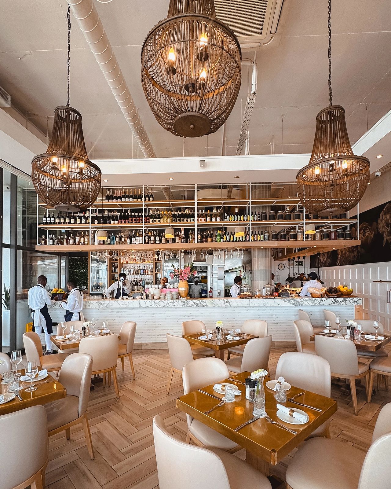 Bright and elegant restaurant interior with warm wooden tones, tan leather chairs, and gold-accented tables neatly set for service. Large woven chandeliers hang from the ceiling above a marble bar backed by shelves filled with bottles and jars. Waitstaff in white shirts and navy aprons prepare drinks while guests begin to arrive. The space has a sophisticated, modern coastal vibe. BLily's elegant interior with woven chandeliers, marble bar, and cream leather seating in Mouille Point, Cape Town