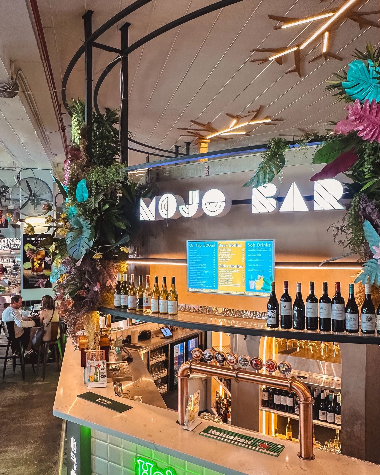 Mojo Market bar interior with tropical decor and neon signage in Sea Point. Text reads: "MOJO BAR," menu boards with drink selections