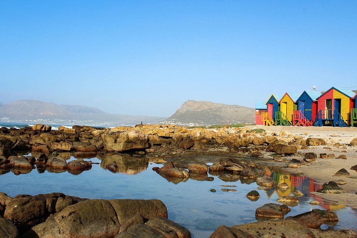 Colorful wooden beach huts in red, yellow, blue, and green line the sandy shore of Muizenberg Beach in Cape Town. Rocky tidal pools and large boulders fill the foreground, reflecting the bright huts in the still water. In the background, mountains rise under a clear blue sky, creating a striking contrast with the vibrant structures. Colorful Muizenberg beach huts reflected in tidal pools with Table Mountain across False Bay, surf culture in Cape Town guide