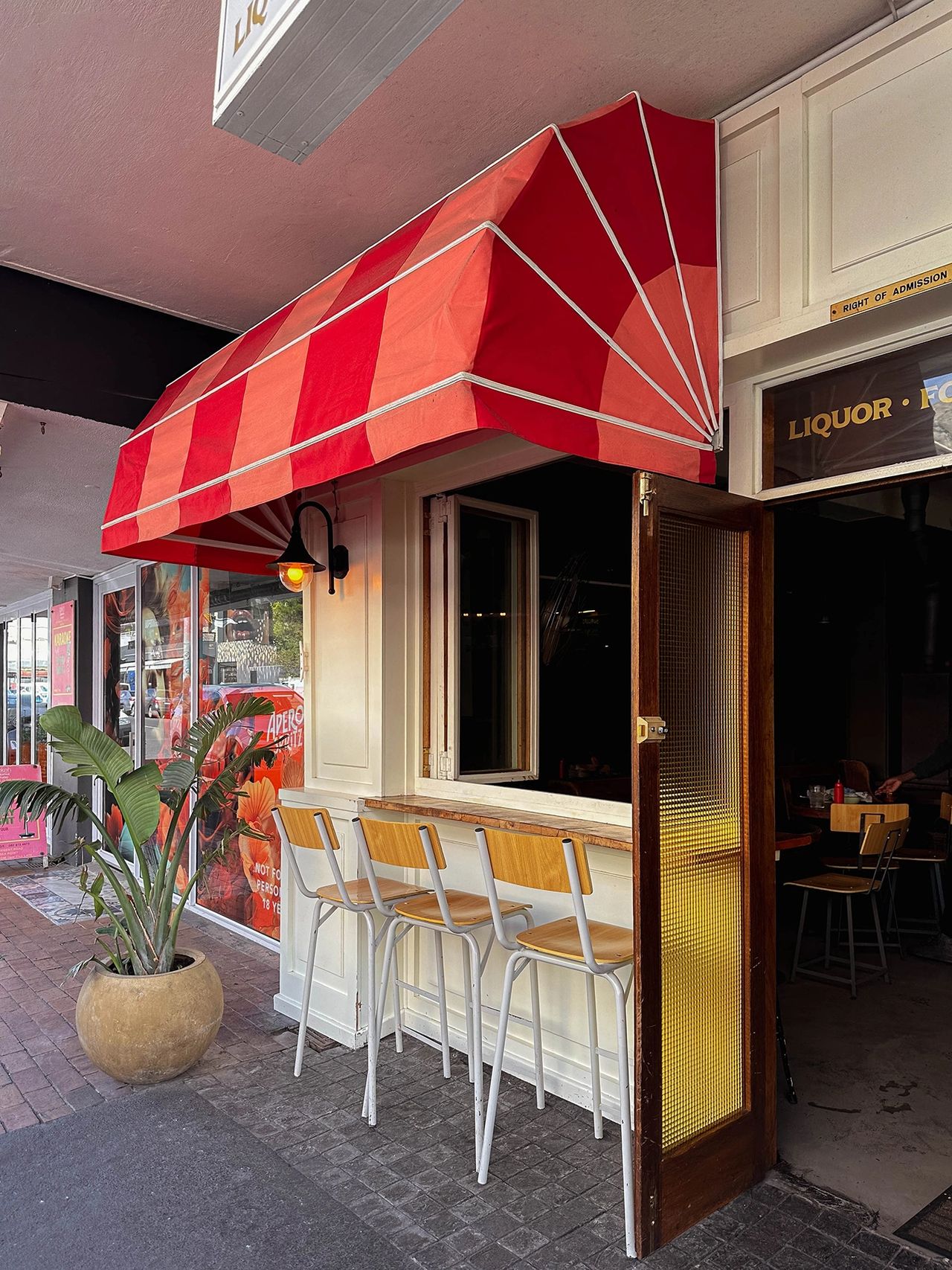 Street view of a cozy bar or café with a bold red-and-pink striped awning and three wooden bar stools lined up at a window counter. A potted tropical plant sits nearby, and the door to the interior is propped open, offering a peek inside. The space has a retro, welcoming vibe ideal for casual drinks or people-watching. Only Fools in Sea Point, Cape Town Lily's restaurant exterior with red striped awning and sidewalk seating on Mouille Point promenade, Cape Town