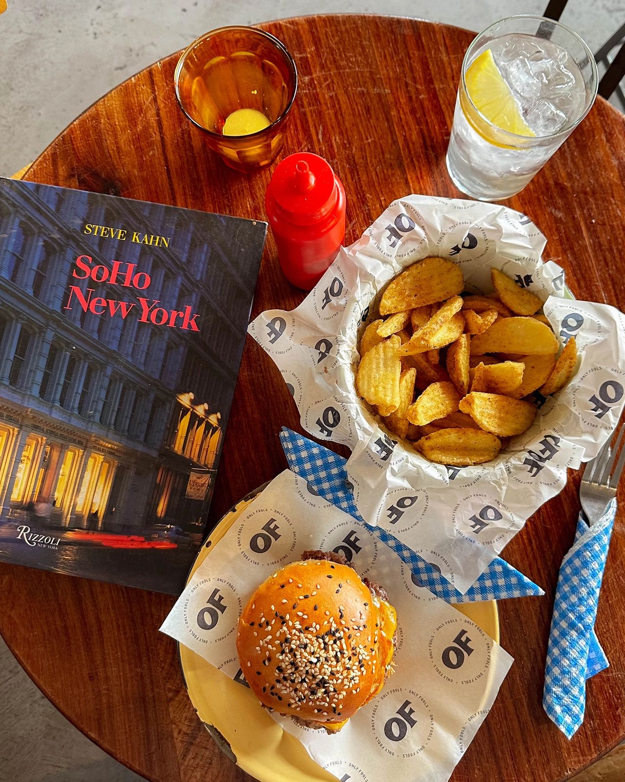 Overhead view of a wooden table set with a sesame seed-topped cheeseburger, a basket of crinkle-cut fries, a red ketchup bottle, a drink with lemon, and a yellow cocktail. A blue checkered napkin and a book titled SoHo New York by Steve Kahn complete the cozy, casual dining setup at Only Fools, a trendy burger spot. Gourmet burger with sesame bun and seasoned chips at Only Fools Cape Town, alongside SoHo New York coffee table book