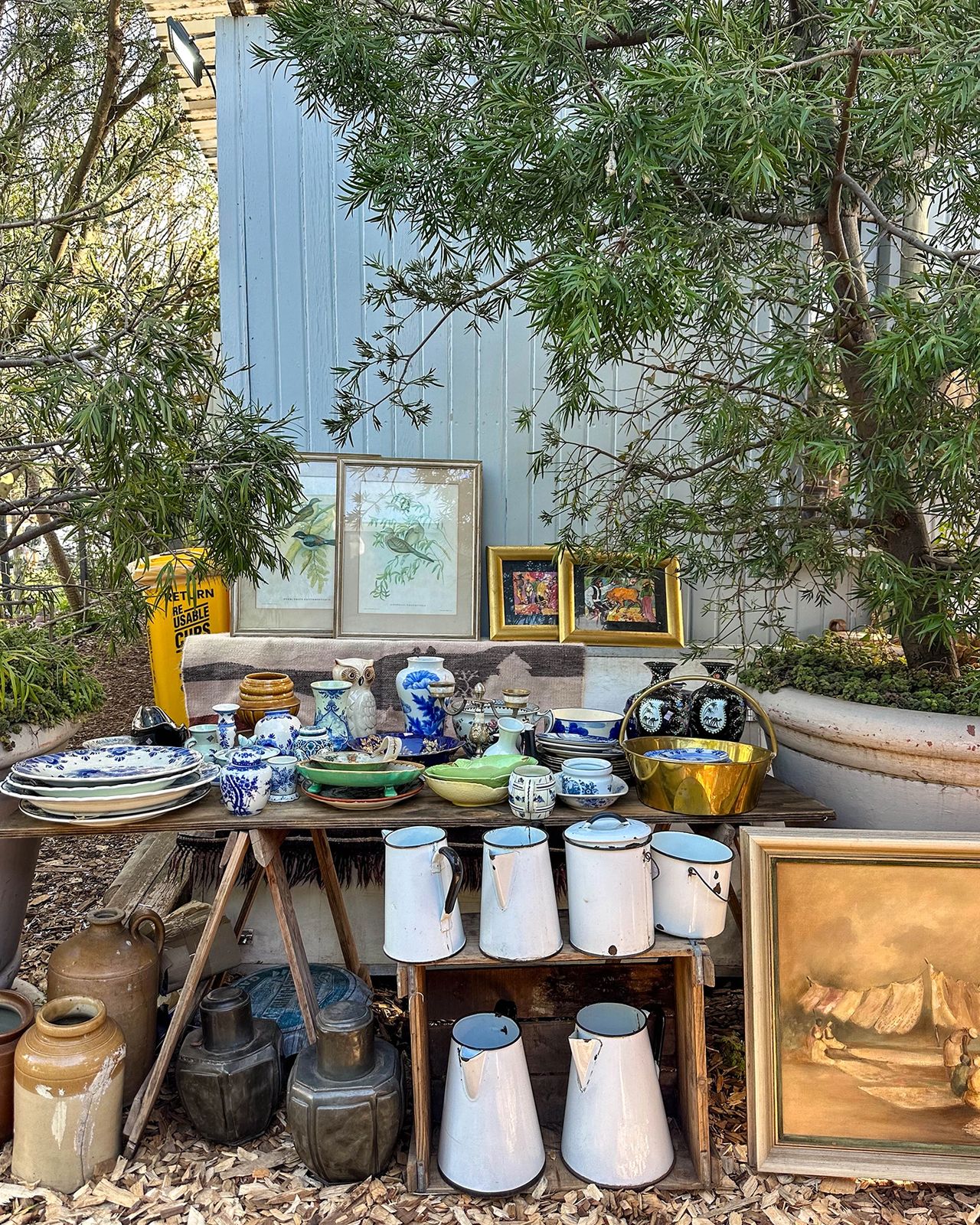 Outdoor display of vintage ceramics, jugs, and framed artwork at a market stall. The table is filled with blue-and-white porcelain, rustic kitchenware, and antique treasures set beneath leafy trees and next to a light blue building. Vintage enamelware, blue pottery, and antique paintings at Oranjezicht City Farm Market, weekend treasure hunting in Cape Town