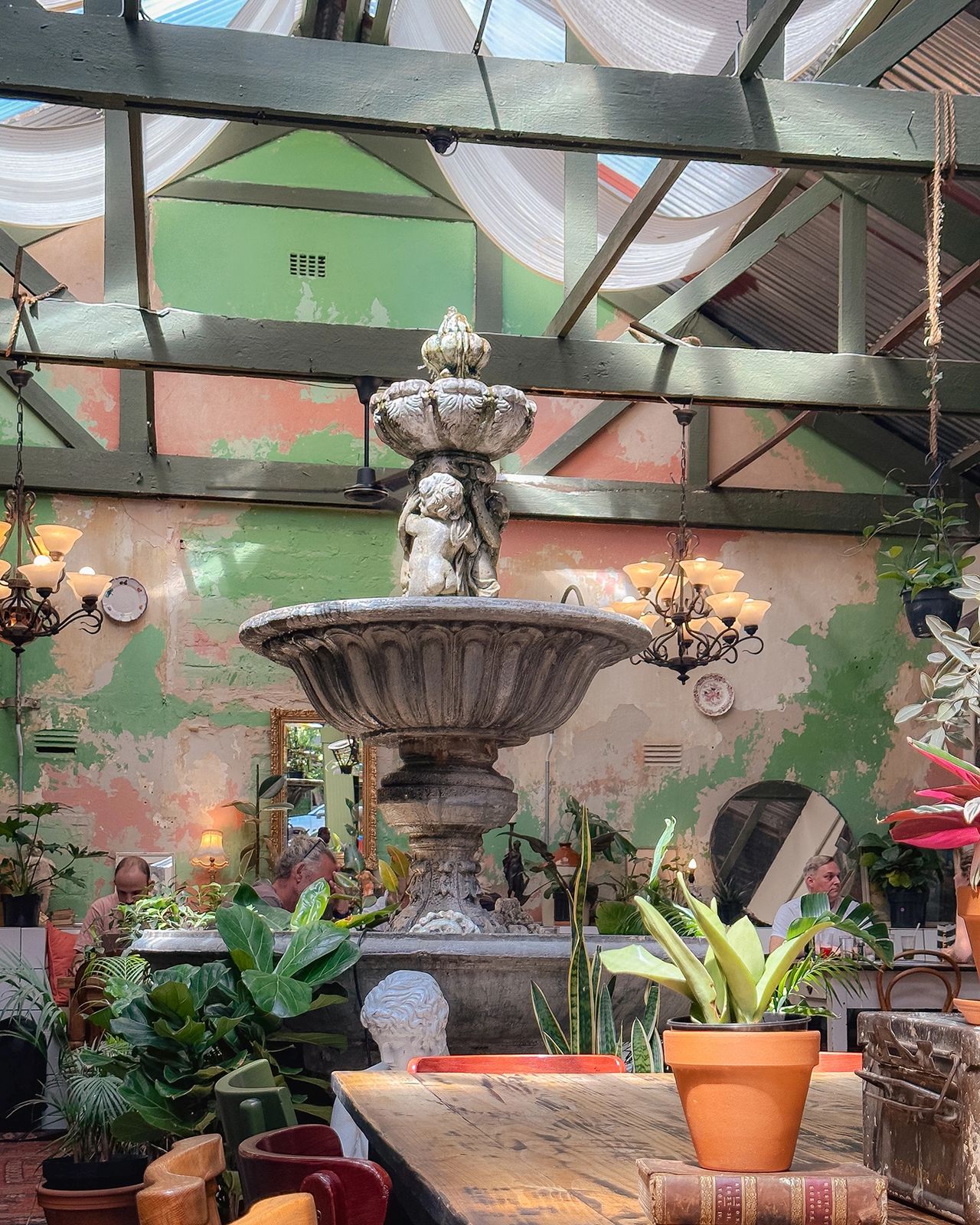 The image shows a charming café interior with a large vintage fountain as the centerpiece. Surrounded by potted plants, antique chandeliers, rustic wooden tables, and distressed green walls, the space has a whimsical, garden-like feel. Natural light pours in from above, creating a warm, inviting atmosphere. TOur Local café garden room with European stone fountain, hanging plants, and eclectic vintage décor in Cape Town