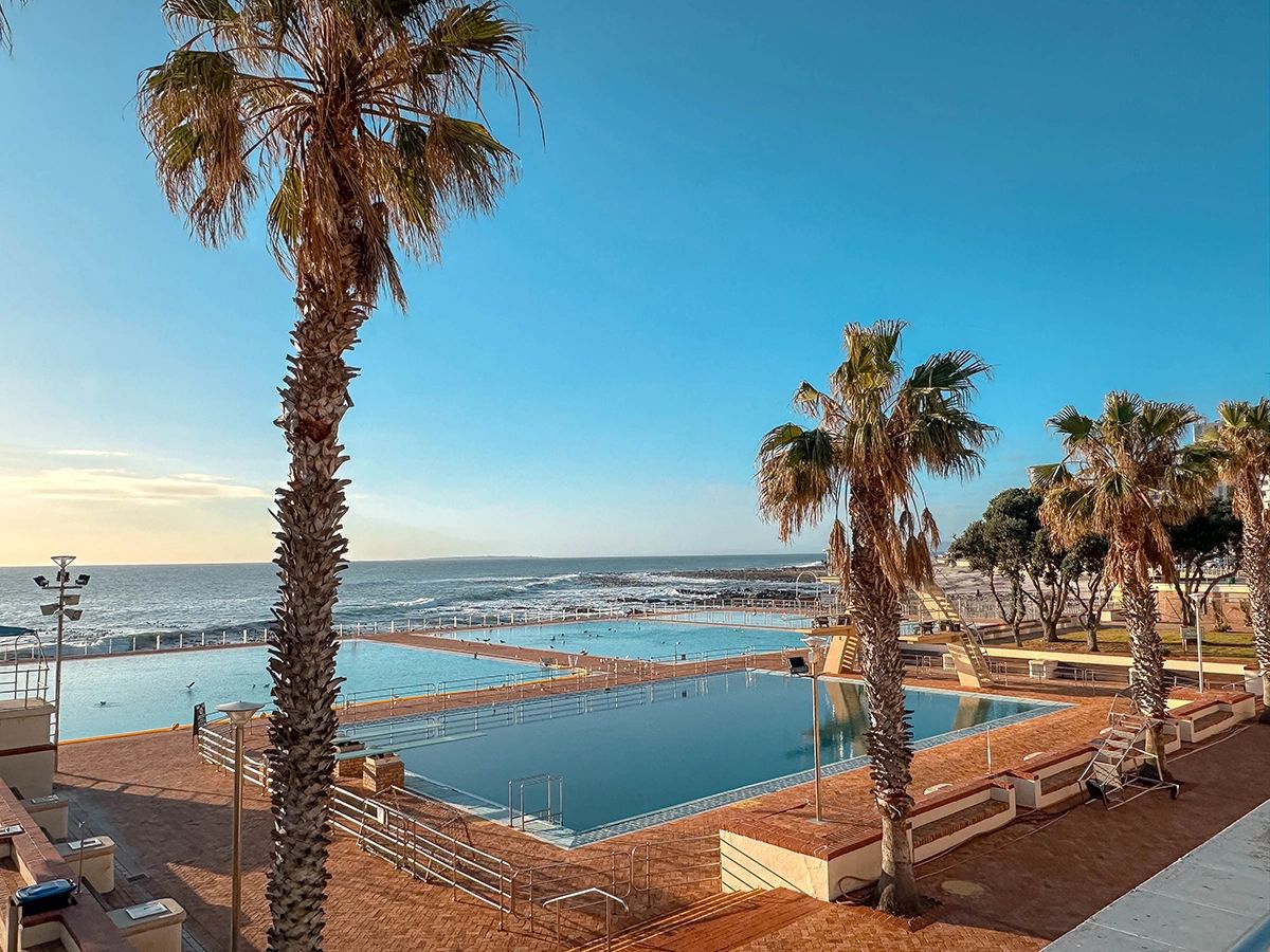 This sunny image showcases the Sea Point Pavilion Pools in Cape Town—an oceanfront public pool complex that offers crystal-clear saltwater pools just steps from the Atlantic. Located along the Sea Point Promenade, it’s a favorite among locals for laps, lounging, and family fun, all with panoramic views of the ocean. Insider tip: arrive early for a peaceful swim, and grab a coffee or bite from one of the nearby cafés post-dip. It’s one of the best-kept gems for cooling off in Cape Town’s warmer months. Sea Point Pavilion public pools with palm trees and Atlantic Ocean backdrop, local favorite in this Cape Town guide