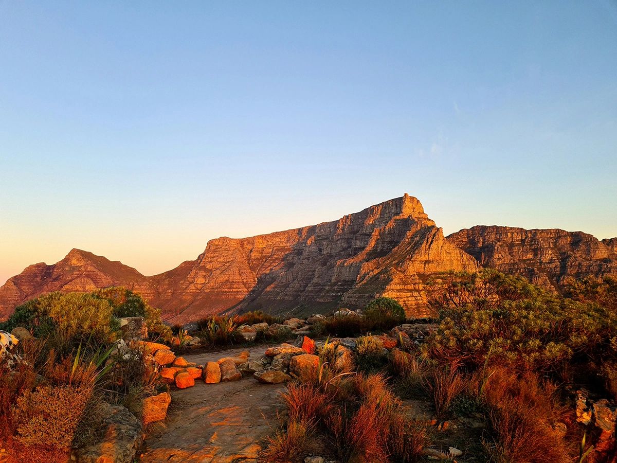 This breathtaking photo captures Table Mountain glowing in golden light during sunset—one of the most iconic and awe-inspiring views in Cape Town, South Africa. Whether you hike to the summit or take the cable car up, the panoramic vistas from the top are truly unforgettable. Surrounded by endemic fynbos and dramatic cliffs, Table Mountain is a must-visit for nature lovers, photographers, and anyone looking to experience the natural beauty of the city from above. Golden sunset at Table Mountain summit with hikers on rocky trail and fynbos vegetation, iconic Cape Town guide experience