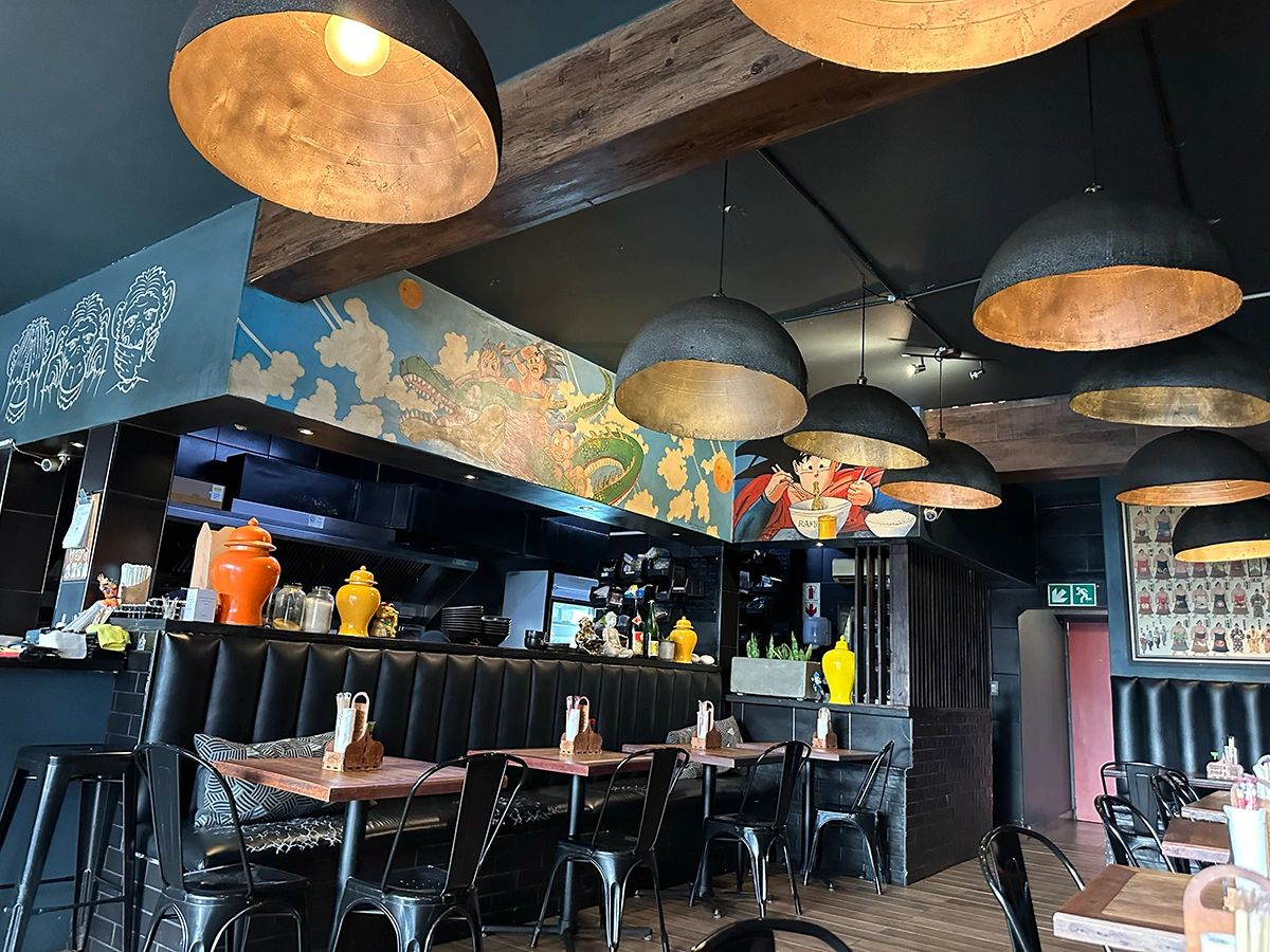 Interior of a trendy Japanese-inspired ramen restaurant with black leather booth seating, wooden tables, and industrial-style black and gold pendant lights. Colorful murals, including a cartoon character eating ramen and a dragon in the clouds, decorate the walls above the open kitchen. The space has a playful, modern, and cozy atmosphere. Three Wise Monkeys ramen bar interior with Japanese murals, industrial pendant lights, and colorful temple jars in Sea Point