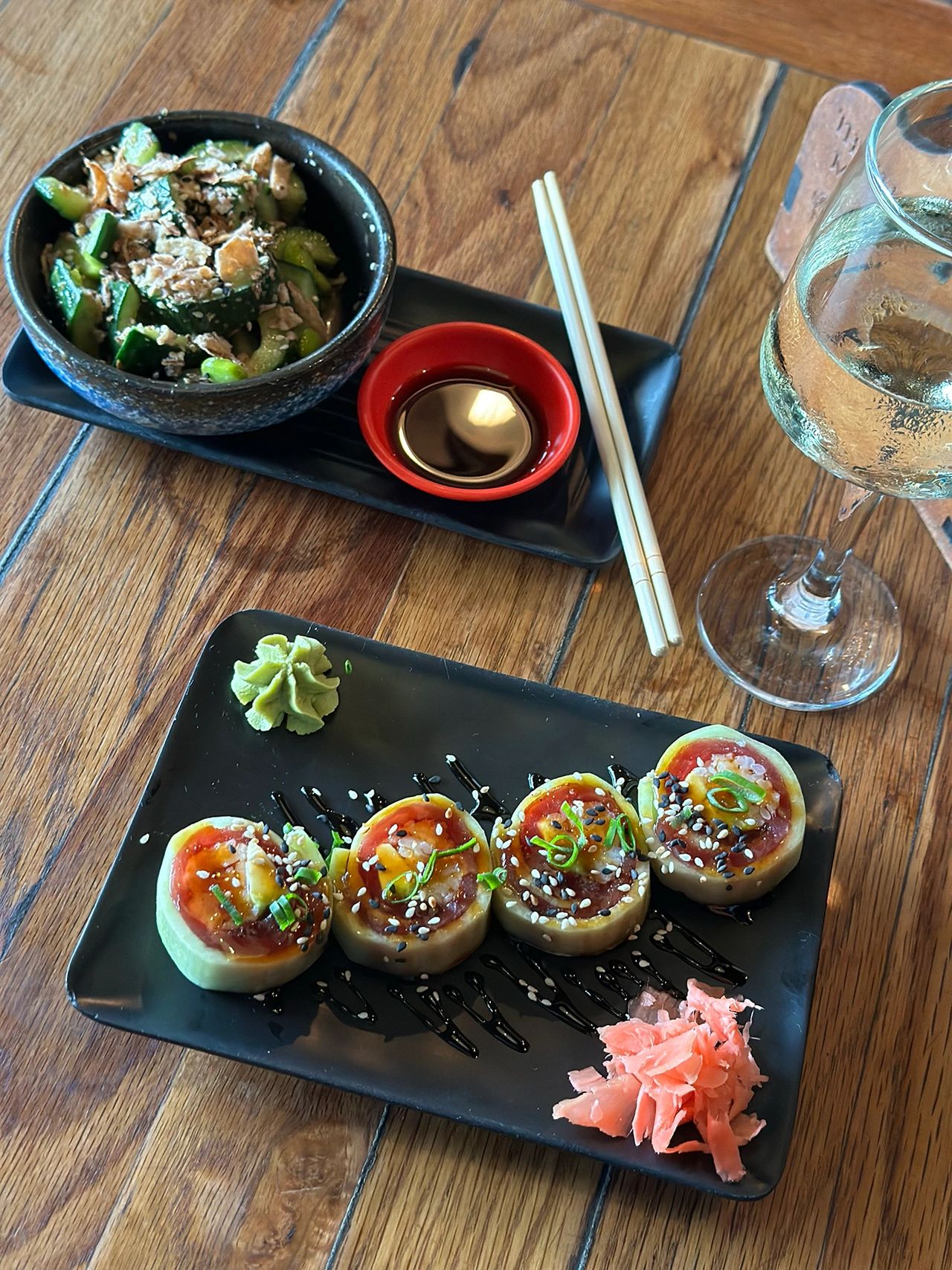 Close-up of a beautifully plated Japanese meal on a wooden table. The black rectangular plate holds five pieces of cucumber-wrapped sushi topped with sauce, sesame seeds, and scallions, alongside wasabi and pickled ginger. A bowl of cucumber salad with shredded fish and a small dish of soy sauce sit nearby, with chopsticks and a glass of white wine completing the elegant dining setup. Fresh sushi rolls with cucumber salad, white wine, and chopsticks at Three Wise Monkeys, a local favorite in this Cape Town guide